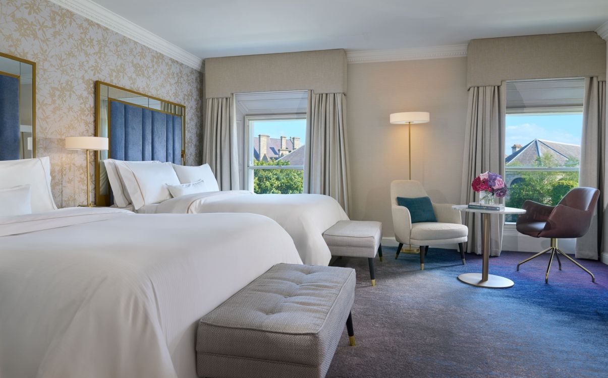 Two double beds with blue headboard and carpet, large windows with view of city in The College Green Hotel Dublin